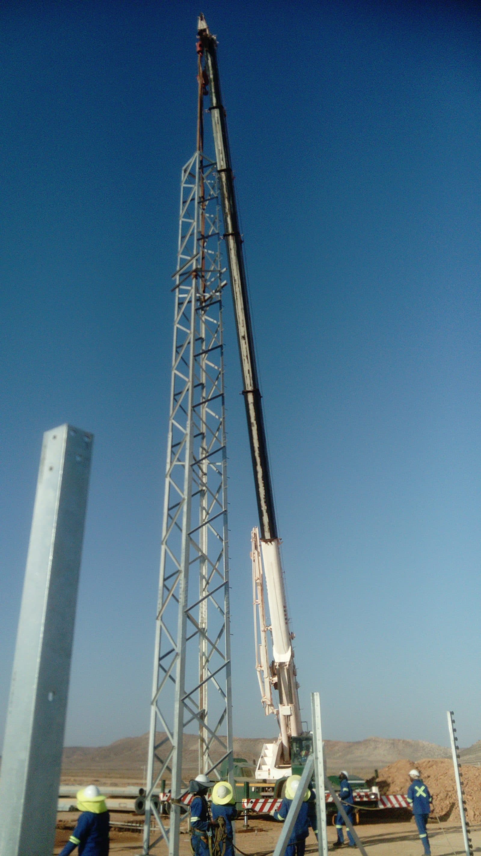 A crane lifting a steel mast into position while crew members in PPE guide it