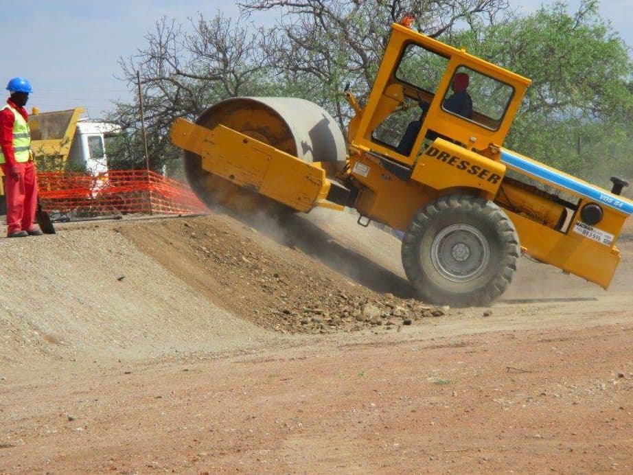 A roller compacting the substation platform while a flagman in PPE keeps watch